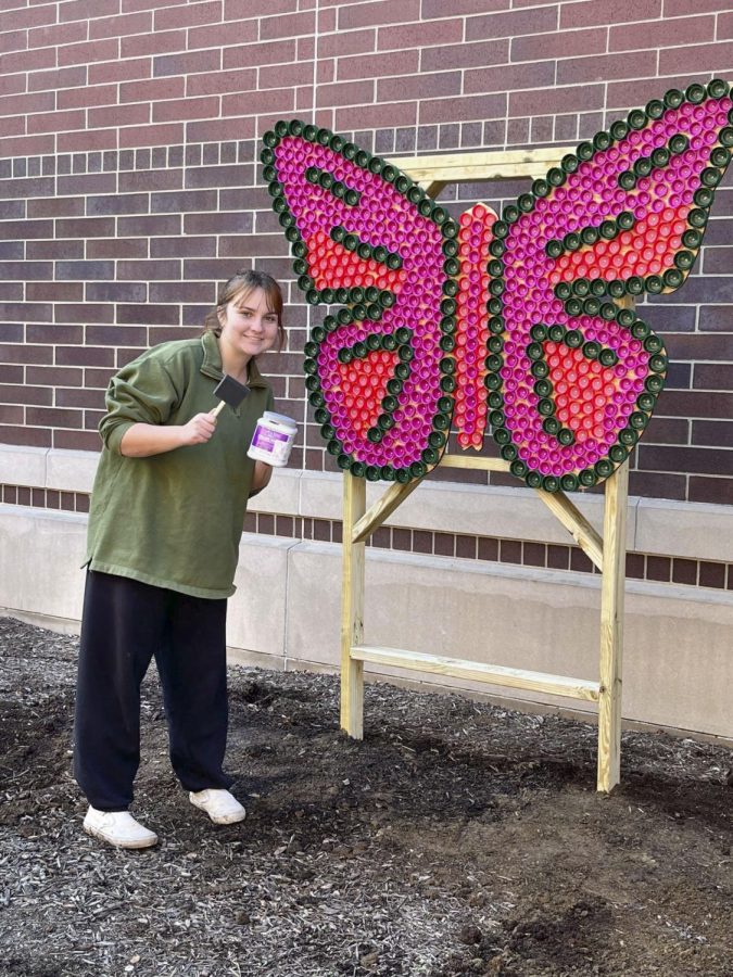 Senior Kaelyn Hart poses in front of her handmade butterfly sculpture located in the main courtyard. She holds a paint can and brush, ready to add finishing touches to the stand.